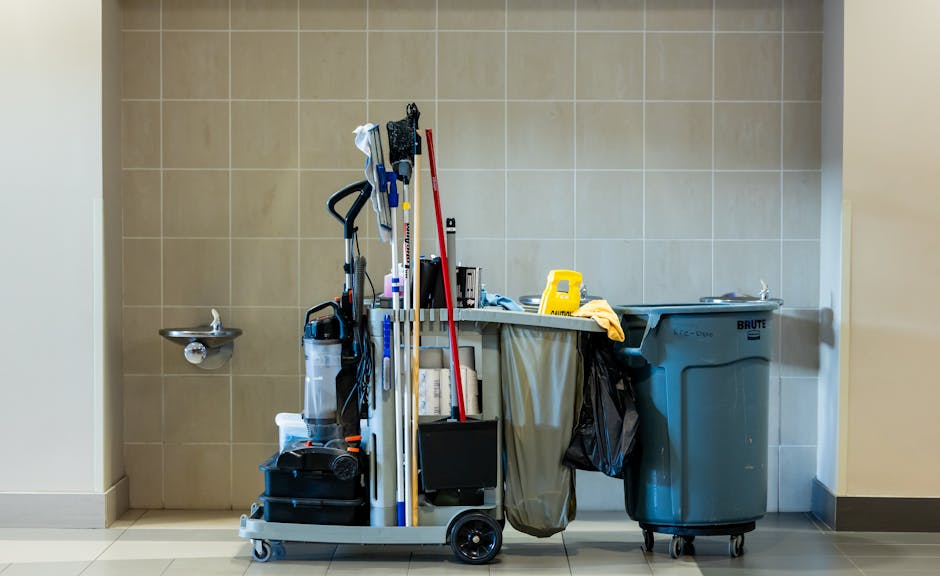 A cleaning cart positioned against a beige tiled wall in a commercial or residential space, with various cleaning tools and supplies including mops, brooms, and spray bottles visible. To the right is
